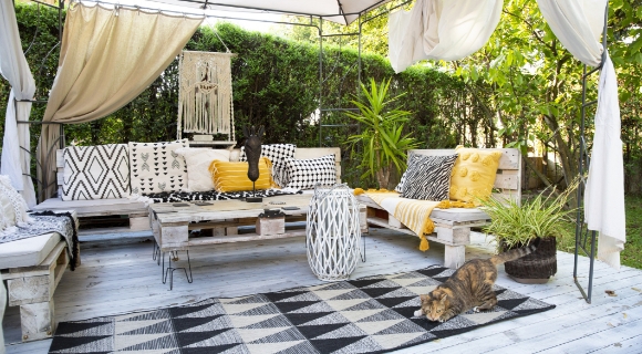 &Uuml;berdachte Gartenlounge mit wei&szlig;en Vorh&auml;ngen, Sitzm&ouml;beln aus Paletten mit gemusterten Kissen, gelben Decken und schwarz-wei&szlig;em Teppich. 