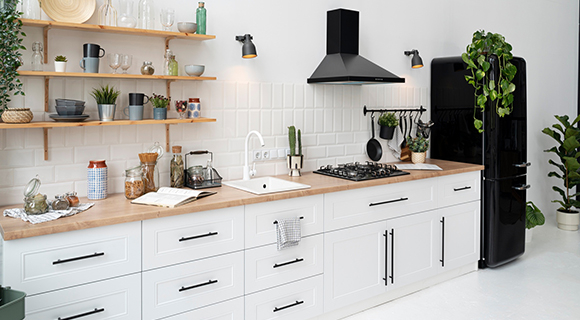 Helle Küche im klassischen Stil mit weißen Fronten, Holzarbeitsplatte, offenen Regalen und schwarzem Retro-Kühlschrank; dekoriert mit grünen Pflanzen und stilvollen Küchenaccessoires.