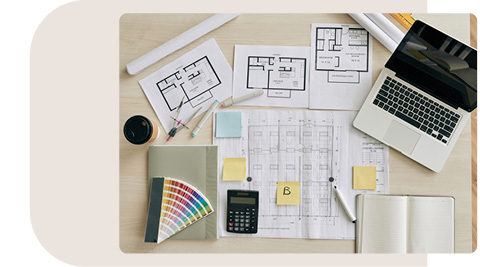 Arbeitsfläche mit Architekturplänen und Arbeitsutensilien. Auf dem Tisch liegen verschiedene Grundrisszeichnungen, ein Farbfächer, ein Taschenrechner, ein Laptop sowie Notizblöcke und Stifte. Post-it-Notizen und Farbmarker ergänzen das kreative Arbeitsumfeld