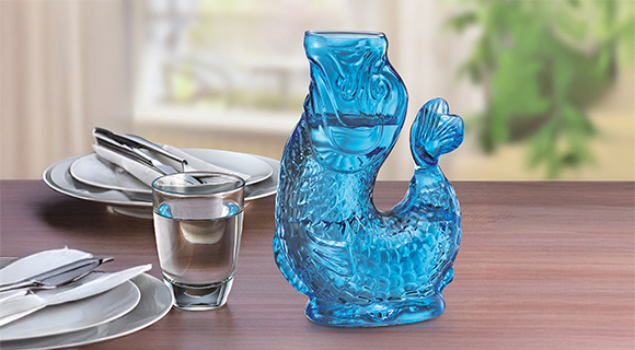 Eine auffällige Vase oder Karaffe aus blauem Glas in Fischform steht auf einem Holztisch. Daneben sind ein Glas Wasser und gedecktes Geschirr arrangiert.