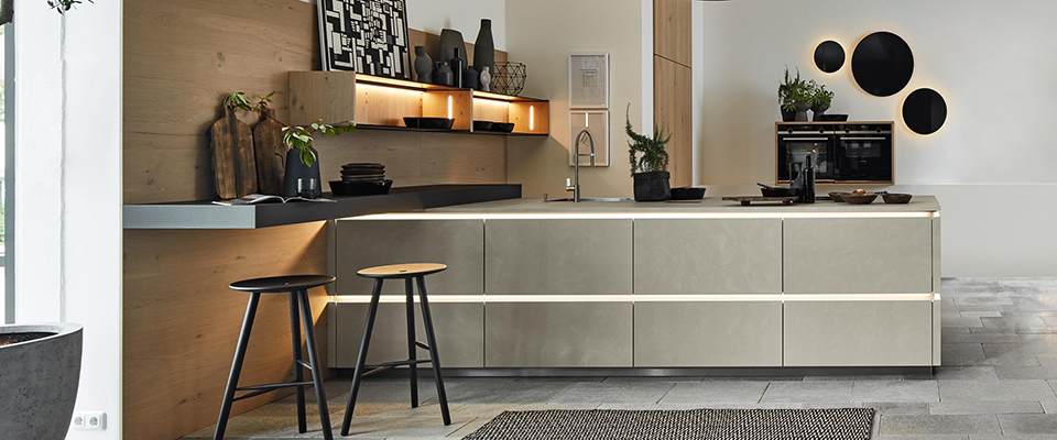 Moderne Designküche mit Kücheninsel in Betonoptik und integrierter LED-Lichtleiste. Die Fronten der Insel sind grifflos und in einem hellgrauen, matten Beton-Look gehalten. Links befindet sich eine Barplatte mit zwei schwarzen Hockern vor einer Wandverkleidung in warmem Holzton, an der offen beleuchtete Regalböden mit schwarzen Vasen, Geschirr und Dekorationen angebracht sind. Rechts in der Wand sind moderne Einbaugeräte integriert. Darüber hängen drei runde Wanddekorationen in Schwarz mit indirekter Beleuchtung. Der Boden ist aus großformatigen grauen Steinfliesen, was der Küche einen eleganten, urbanen Charakter verleiht. Eine dunkel gemusterte Fußmatte liegt im Vordergrund. Die gesamte Atmosphäre wirkt stilvoll, hochwertig und architektonisch durchdacht.