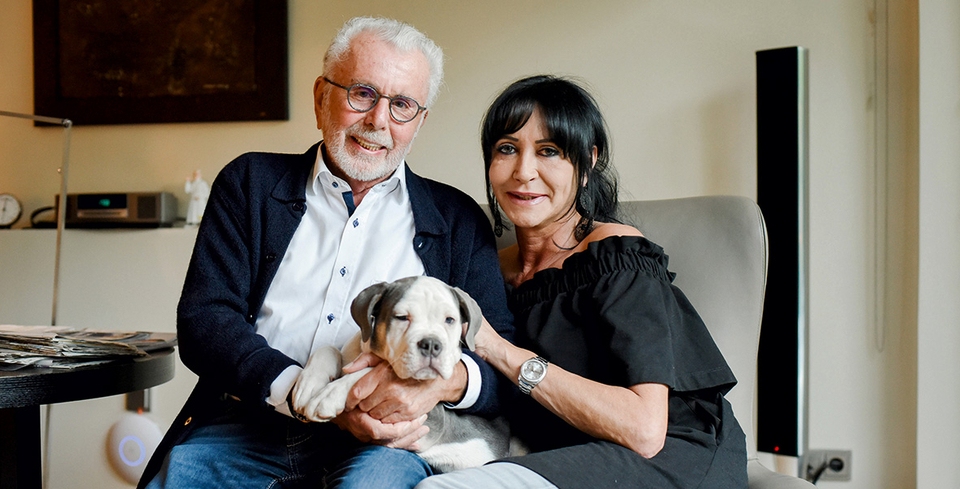 Das Bild zeigt ein &auml;lteres Paar, das gemeinsam auf einem Sitzm&ouml;bel in einem Wohnraum sitzt. Zwischen ihnen halten sie einen jungen Hund auf dem Scho&szlig;. Die Personen blicken in Richtung Kamera und wirken entspannt. Im Hintergrund sind Wohnaccessoires und technische Ger&auml;te zu erkennen, die auf eine private Wohnumgebung hinweisen.