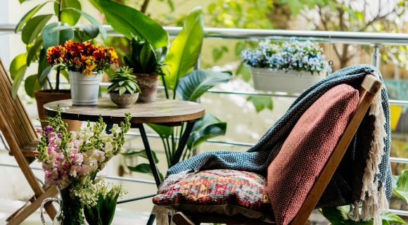 Gartenklappstuhl mit bunten Outdoorkissen steht auf einem Balkon, der mit verschiedenen Blumen und Pflanzen verziert ist.