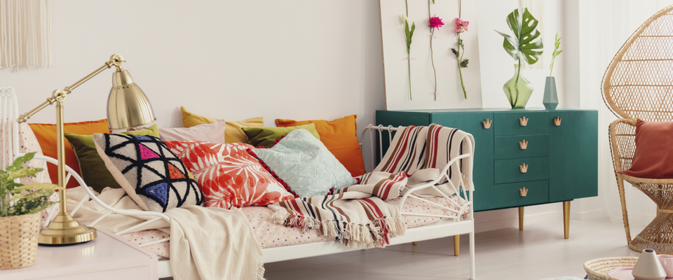 Boho-Wohnzimmer mit bunter Kissenlandschaft, Rattansessel, grüner Kommode und floralen Deko-Elementen