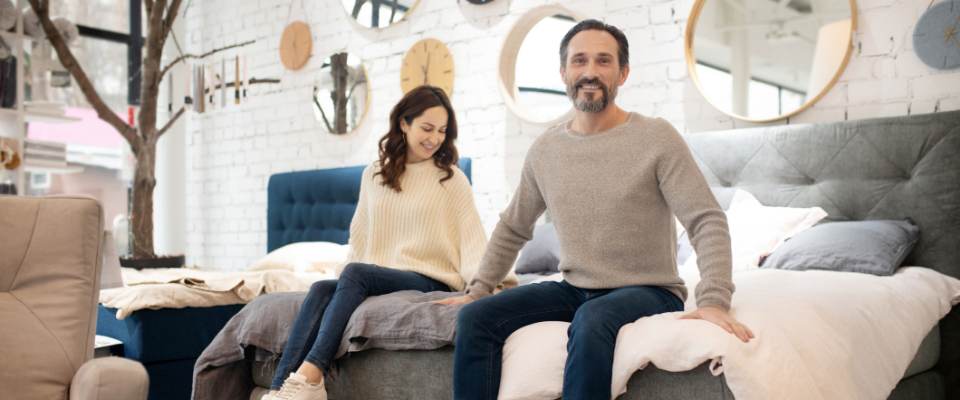 Paar sitzt auf einem Boxspringbett im Einrichtungshaus und testet Sitzhöhe und Komfort – stilvolle Umgebung mit modernen Spiegeln und gemütlicher Atmosphäre