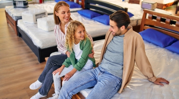 Ein Elternpaar sitzt mit ihrer Tochter auf einer Matratze in einem Möbelhaus und lächelt sich an.