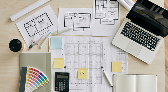 Schreibtisch mit Unterlagen zur Küchen- oder Wohnraumplanung, darunter Baupläne, Grundrisse, Farbfächer, Laptop, Notizbuch und weitere Arbeitsmaterialien wie Stifte, Taschenrechner und Haftnotizen.