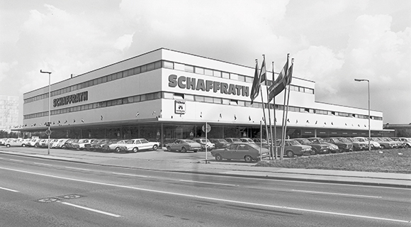 Das Bild zeigt ein historisches Schwarzwei&szlig;foto eines gro&szlig;en M&ouml;belhauses mit der Aufschrift &bdquo;Schaffrath&ldquo;. Vor dem mehrst&ouml;ckigen Geb&auml;ude befindet sich ein Parkplatz mit zahlreichen abgestellten Autos sowie eine mehrspurige Stra&szlig;e im Vordergrund. Mehrere Fahnenmasten stehen vor dem Eingangsbereich des Geb&auml;udes. Die Aufnahme vermittelt einen Eindruck der damaligen Architektur und des Umfelds des Wohnkaufhauses.