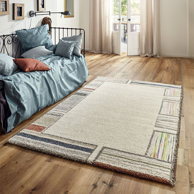 Hochflorteppich mit cremefarbener Fl&auml;che und gemustertem Rand in bunten Farben liegt auf einem Holzboden in einem hellen Wohnzimmer. Im Hintergrund steht ein schwarzes Metallsofa mit blauer Tagesdecke und mehreren Kissen. Helle Vorh&auml;nge rahmen eine ge&ouml;ffnete Doppelt&uuml;r mit Blick ins Freie.