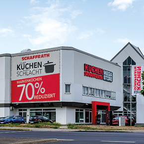 Das Bild zeigt die Au&szlig;enansicht eines K&uuml;chenfachmarkts mit dem Schriftzug &bdquo;K&uuml;chen Schaffrath Mega Store&ldquo; an der Fassade. An der Geb&auml;udeseite ist ein gro&szlig;fl&auml;chiges Banner mit Hinweisen zu einer K&uuml;chenaktion angebracht. Der Eingangsbereich ist durch farblich abgesetzte Elemente deutlich erkennbar. Vor dem Geb&auml;ude verlaufen eine Stra&szlig;e sowie angrenzende Gr&uuml;nfl&auml;chen.