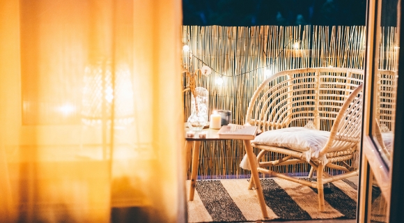 Balkonecke mit Rattanstuhl, kleinem  Holztisch mit Kerze und Buch sowie Lichterkette  vor Bambus-Sichtschutz bei Abendlicht