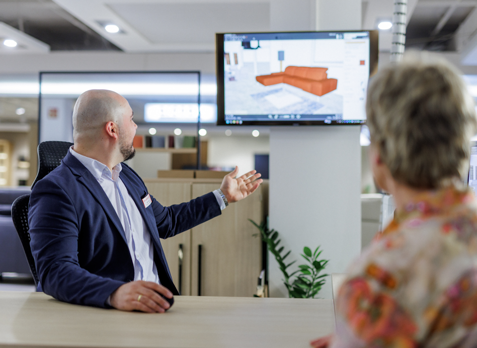 Zwei Personen sitzen w&auml;hrend einer M&ouml;belberatung an einem Tisch, im Hintergrund sieht man einen Bildschirm, auf dem ein Sofa via 3-D Planungstool in einen Raum eingef&uuml;gt wird.