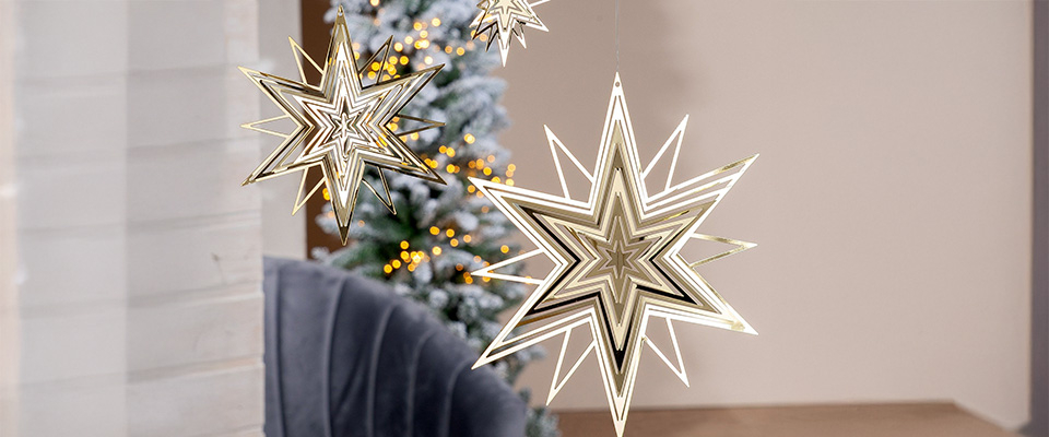 Mehrere goldene Leuchtsterne hängen vor einem weihnachtlich geschmückten Baum. Das warme Licht erzeugt eine festliche und elegante Stimmung.