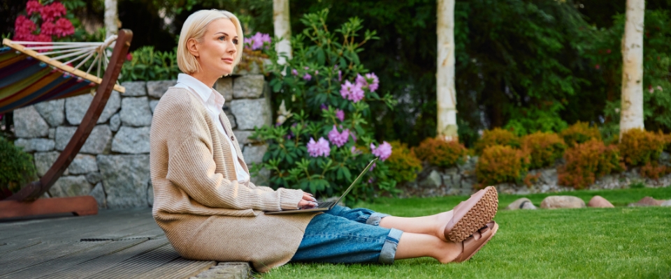 Person sitzt mit einem Laptop auf einer Rasenfläche im Garten, neben einer Steinmauer und blühenden Sträuchern. 