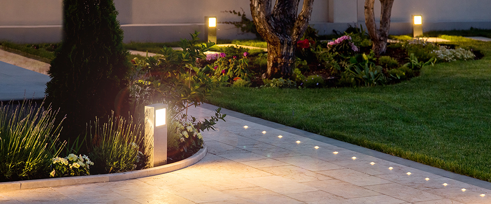 Beleuchteter Gartenweg mit eingelassenen Bodenleuchten und modernen Pollerleuchten entlang gepflegter Grünflächen.