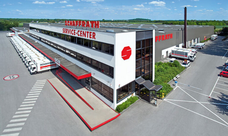Das Bild zeigt das „Schaffrath Service-Center“, ein großes, modernes Logistik- und Lagergebäude. Die Fassade des Gebäudes ist weiß mit großen Fensterfronten und roten Akzenten. Auf dem Dach steht in großen roten Buchstaben der Schriftzug „SCHAFFRATH SERVICE-CENTER“. Auf der linken Seite sind zahlreiche Laderampen zu sehen, an denen mehrere Schaffrath-LKWs geparkt sind. Der Eingangsbereich ist mit einem roten Vordach versehen und führt in ein gläsernes Foyer. Rechts vom Gebäude erstreckt sich ein Parkplatz mit Autos, Grünflächen und Bäumen, die das industrielle Erscheinungsbild auflockern. Im Hintergrund sind Felder und ein blauer Himmel mit wenigen weißen Wolken sichtbar, was dem Bild eine helle und offene Atmosphäre verleiht.