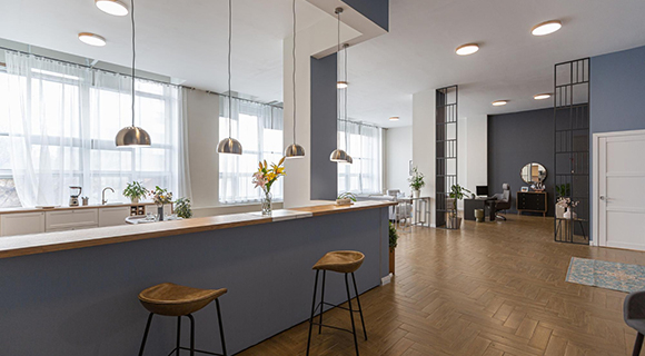 Großzügige, modern eingerichtete Büroküche mit freistehender Kochinsel in Blau und einer hellen Holzarbeitsplatte. Vor der Insel stehen zwei Barhocker mit braunen Ledersitzen und schwarzem Metallgestell. Darüber hängen vier silberne Pendelleuchten. Die Rückwand der Küche ist mit weißen Unterschränken, Spüle, Pflanzen und Vasen dekoriert. Der Raum ist lichtdurchflutet durch große Fenster mit weißen Vorhängen. Rechts im Hintergrund befindet sich ein Bürobereich mit Schreibtischen, Pflanzen, Teppich und dekorativen Raumteilern in Schwarz. Die Atmosphäre wirkt offen, freundlich und wohnlich – ideal für gemeinsames Arbeiten und Pausen in entspannter Umgebung.