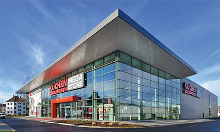 Das Bild zeigt den „Küchen Schaffrath Mega Store“, ein modernes Gebäude mit einer beeindruckenden Glasfassade und einem flachen, überstehenden Dach aus Metall. Die Glasfront spiegelt den blauen Himmel und sorgt für ein lichtdurchflutetes Erscheinungsbild. Auf der rechten Seite der Fassade befindet sich der Schriftzug „KÜCHEN SCHAFFRATH MEGA STORE“ in großen, roten und weißen Buchstaben. Der Eingangsbereich wird durch einen roten Rahmen hervorgehoben, der das Design modern und einladend macht. Links neben der Glasfront ist ein großes Werbebanner mit Küchenmotiven zu sehen. Der Himmel ist klar und strahlend blau, was das Gebäude in einem freundlichen Licht präsentiert.