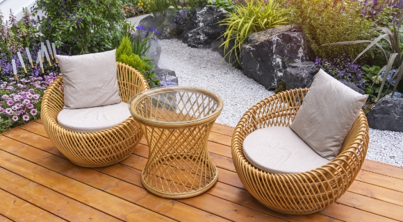 Zwei runde Rattan-Loungesessel mit hellen Sitzkissen und kleiner runder Beistelltisch auf Holzterrasse neben Kiesbeet und Zierpflanzen.