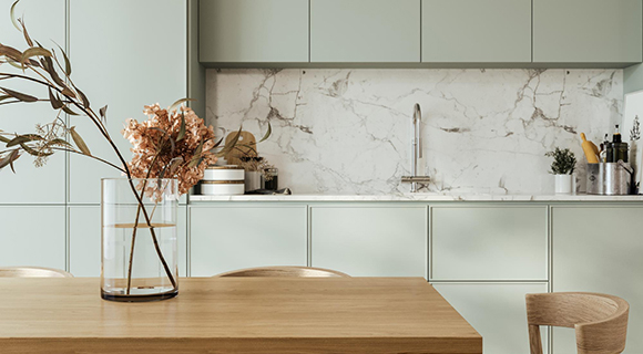 Helle, moderne Küche mit grifflosen, mattgrünen Fronten und einer auffälligen Rückwand aus weißem Marmor mit grauer Maserung. Im Mittelpunkt steht ein schlichter Wasserhahn über einer eingebauten Spüle. Die Ober- und Unterschränke sind klar strukturiert und fügen sich nahtlos in das reduzierte Design ein. Im Vordergrund steht ein Esstisch aus hellem Holz mit passenden Stühlen. Auf dem Tisch befindet sich eine hohe Glasvase mit dekorativen Trockenblumen in warmen Naturtönen. Die Küche wirkt aufgeräumt, stilvoll und von natürlichem Licht durchflutet.