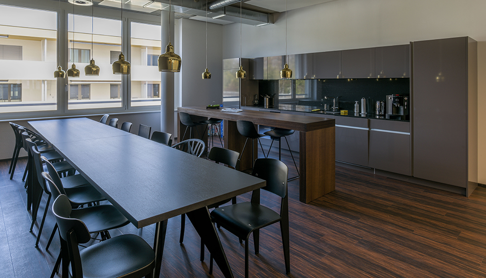 Große, modern gestaltete Büroküche mit dunklem Holzfußboden und grauen, hochglänzenden Fronten. Die Küchenzeile im Hintergrund erstreckt sich über die gesamte Wand und enthält integrierte Geräte wie Wasserkocher, Toaster und eine Kaffeemaschine. Davor steht ein hoher Holztresen mit vier schwarzen Barhockern, ideal für kurze Pausen. Im Vordergrund befindet sich ein langer Esstisch mit zahlreichen schwarzen Stühlen – geeignet für Teammeetings oder gemeinsame Mittagessen. Mehrere goldfarbene Pendelleuchten hängen über Tresen und Tisch und setzen stilvolle Akzente. Durch große Fenster fällt viel Tageslicht in den Raum. Die Atmosphäre wirkt hochwertig, offen und funktional – perfekt für moderne Arbeitsumgebungen.