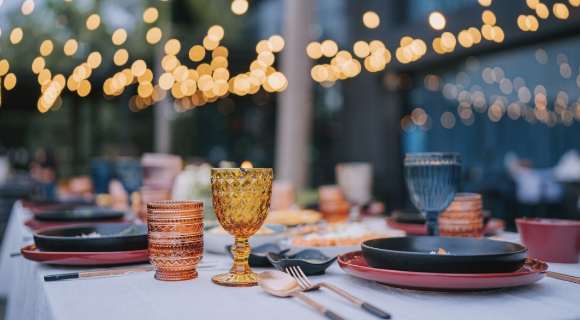 Gedeckter Outdoor-Esstisch mit bunten Gl&auml;sern, Tellern und Besteck, im Hintergrund warm leuchtende Lichterketten in Unsch&auml;rfe.