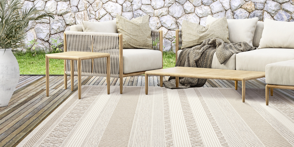 Gemütliche Terrasse mit hellen Loungemöbeln, Holztischen und beigem Outdoor-Teppich auf Holzdeck vor Natursteinmauer.