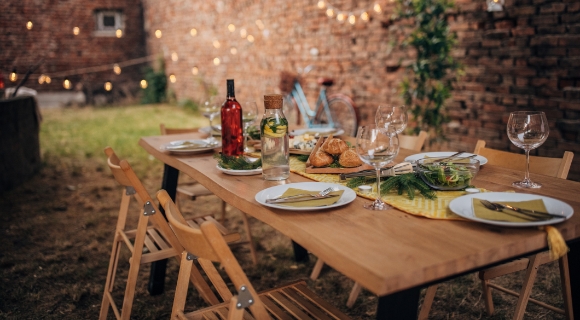 Gedeckter Holztisch im Garten mit Tellern, Gl&auml;sern, Brot und Lichterkette vor Ziegelwand.