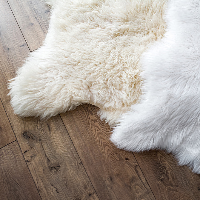 Zwei flauschige Teppiche in Felloptik auf dunklem Holzfu&szlig;boden: links ein cremefarbenes Schaffell mit lockiger Struktur, rechts ein glattes, wei&szlig;es Kunstfell mit seidigem Glanz.
