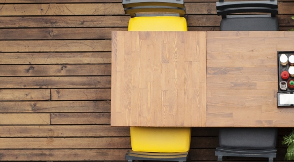 Rechteckiger Holztisch mit gelbem und grauem Stuhl auf einer Terrasse aus Holzbohlen, von oben fotografiert.