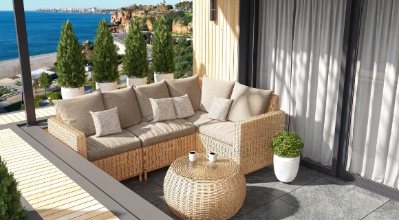 Ecksofa aus Geflecht mit beigen Polstern und rundem Tisch auf Balkon mit Blick auf Meer und K&uuml;stenlandschaft.