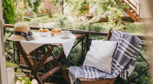 Ein gem&uuml;tlicher Balkon mit einem Holztisch, auf dem eine Schale mit Pfirsichen, ein Kaffeebecher und ein Strohhut liegen. Der Balkon ist von gr&uuml;nen Pflanzen und B&auml;umen umgeben.