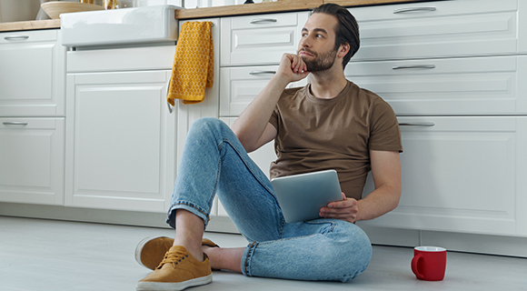 Junger Mann sitzend auf dem Boden einer Einbauküche, in der Hand ein Tablet, im Hintergrund Spülbecken.