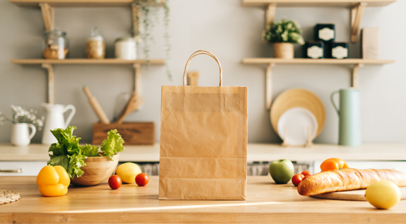 Küche mit Holzregalen im Hintergrund, auf dem Tisch frische Zutaten und eine braune Papiertüte.