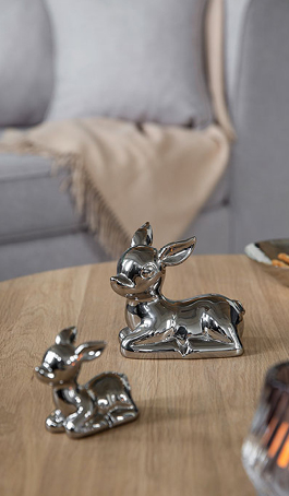 Zwei silberfarbene Dekofiguren in Rehkitzform stehen auf einem Holztisch. Im Hintergrund sorgt eine Decke auf dem Sofa für eine gemütliche Wohnatmosphäre.