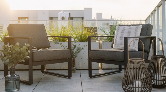 Zwei gepolsterte Loungesessel mit Kissen auf Balkonterrasse neben Pflanzk&uuml;beln und Laternen mit Kerzen