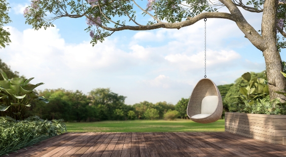 Hängesessel aus Korbmaterial unter einem Baum über einer Holzterrasse mit freier Sicht auf eine Wiese und Pflanzen.