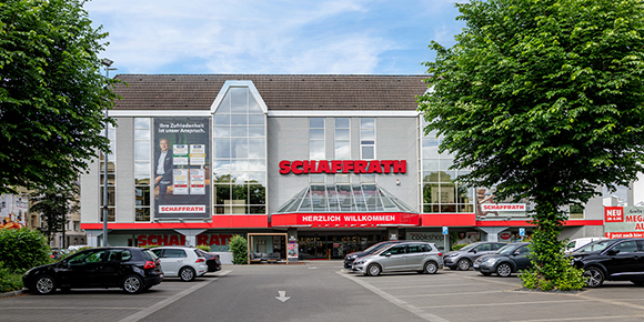 Das Bild zeigt die modernisierte Fassade eines Wohnkaufhauses mit dem Schriftzug &bdquo;Schaffrath&ldquo; &uuml;ber dem Eingangsbereich. Gro&szlig;e Fensterfl&auml;chen erstrecken sich &uuml;ber mehrere Stockwerke und lassen Einblicke in das Geb&auml;ude zu. Vor dem Geb&auml;ude befinden sich parkende Fahrzeuge sowie ein markierter Eingangsbereich mit &Uuml;berdachung. Die Aufnahme zeigt das Geb&auml;ude bei Tageslicht in einer st&auml;dtischen Umgebung.