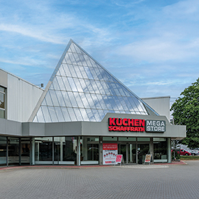 Das Bild zeigt den Eingangsbereich eines Wohnkaufhauses mit einer auff&auml;lligen, pyramidenf&ouml;rmigen Glasarchitektur. &Uuml;ber dem Eingang ist der Schriftzug &bdquo;K&uuml;chen Schaffrath Mega Store&ldquo; angebracht. Die Glasfl&auml;chen spiegeln den Himmel und die umliegenden B&auml;ume wider. Vor dem Geb&auml;ude befindet sich ein gepflasterter Vorplatz mit Hinweistafeln im Eingangsbereich.