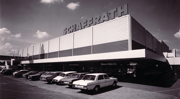 Das Bild zeigt ein historisches Foto eines erweiterten Wohnkaufhauses mit dem Schriftzug &bdquo;Schaffrath&ldquo; auf dem Dach. Vor dem Geb&auml;ude sind mehrere geparkte Pkw aus unterschiedlichen Baujahren zu sehen, die auf einen fr&uuml;heren Aufnahmezeitraum hinweisen. Die Fassade ist horizontal gegliedert und wirkt funktional gestaltet. Der Eingangsbereich befindet sich im Erdgeschoss unter einem auskragenden Geb&auml;udeteil.