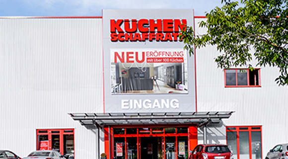 Das Bild zeigt den Eingangsbereich eines K&uuml;chenfachmarkts mit dem Schriftzug &bdquo;K&uuml;chen Schaffrath&ldquo; &uuml;ber dem Haupteingang. An der Fassade sind mehrere gro&szlig;formatige Tafeln mit Hinweisen zu Unternehmenszahlen und Jubil&auml;en angebracht. Ein markierter Fu&szlig;g&auml;nger&uuml;berweg f&uuml;hrt &uuml;ber den Vorplatz direkt zum Eingang. Das Geb&auml;ude ist von bepflanzten Gr&uuml;nfl&auml;chen und einem Parkplatz umgeben.