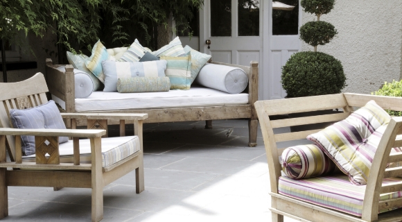 Heller Outdoor-Sitzplatz mit Holzstühlen und gepolsterter Liegebank, umgeben von dekorativen Pflanzen.