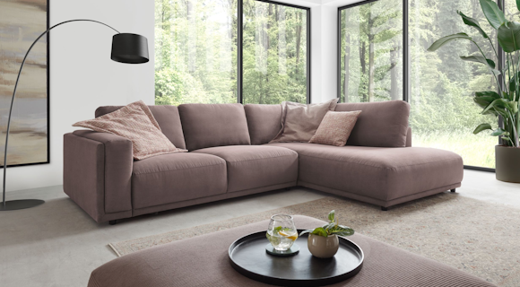 Modernes graues Polstersofa von MONDO steht in einem hellen Wohnzimmer mit gro&szlig;er Fensterfront.