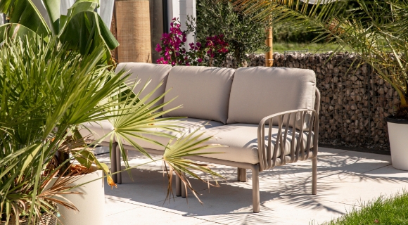 Ecksofa mit hellen Polstern auf gepflasterter Terrasse, umgeben von K&uuml;belpflanzen und Gartenbereich. 