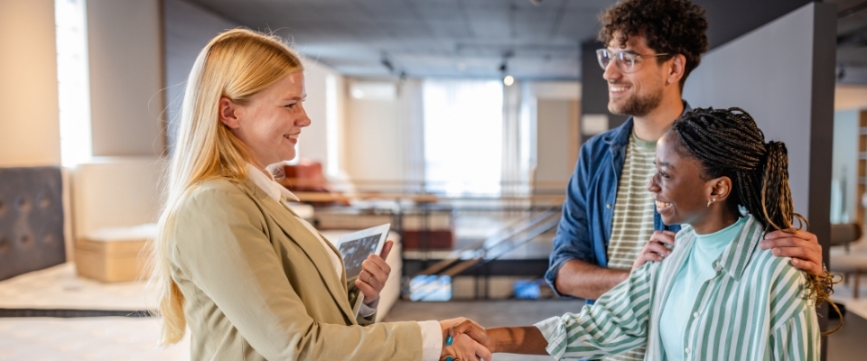 Eine junge Frau gibt einem gegenüberstehendem Pärchen in der Bettenabteilung in einem Möbelhaus die Hand.