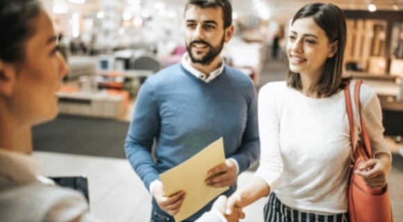 Paar wird freundlich von einer Einrichtungsexpertin im Möbelhaus begrüßt – Start einer persönlichen Wohnberatung