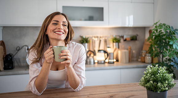 Junge Frau mit Kaffeetasse lehnt an Küchentheke, im Hintergrund helle Einbauküche.