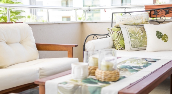 Balkon mit Sofa und Sessel mit hellen Polstern, Holztisch mit Tischl&auml;ufer und dekorativen Kissen.