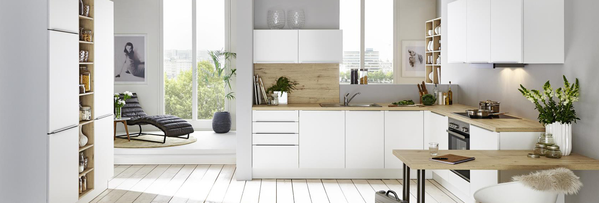 Landhausküche in weiß mit Holzarbeitsflächen und einem gemütlichen, ländlichen Design. Die Küche ist ausgestattet mit Glasschränken, einer Kochinsel und rustikalen Details. Große Fenster im Hintergrund lassen viel Tageslicht herein. Rattan- und Holzmöbel ergänzen den Raum