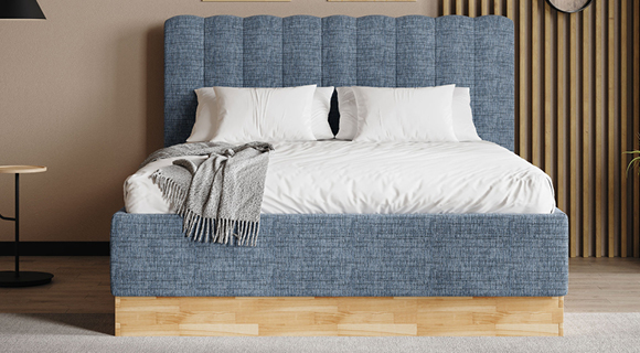 Frontalansicht eines modernen Boxspringbetts mit hohem, weich gepolstertem Kopfteil in jeansblauem Stoff und vertikaler Steppung. Weiße Bettwäsche und mehrere weiße Kissen sorgen für einen klaren, frischen Look; über die linke Bettkante ist eine graue Strickdecke gelegt. Das Bett steht auf einem warmen Holzsockel, der sich vom hellen Teppichboden abhebt. Links ein kleiner runder Beistelltisch mit schwarzer Stehleuchte, rechts eine Wand aus vertikalen Holzlamellen – insgesamt eine ruhige, moderne Schlafzimmeratmosphäre